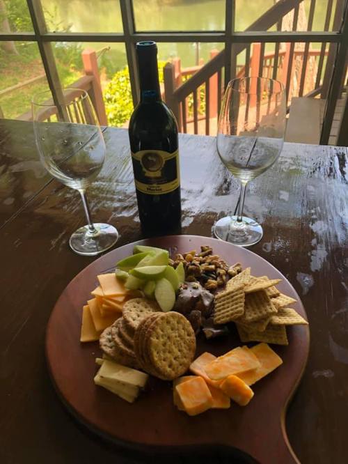 A wooden platter with assorted cheeses, crackers, nuts, and fruit, accompanied by a bottle of wine and two glasses.