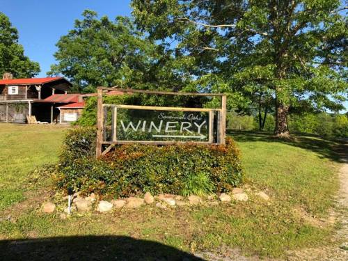 Sign for a winery surrounded by greenery and trees, with a rustic building in the background.
