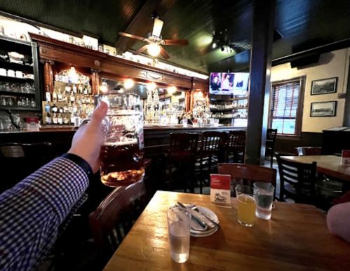 A hand holds a large beer mug in a cozy bar with wooden tables and a well-stocked bar in the background.