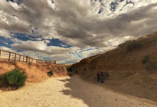 Two people stand in a dry, sandy canyon under a dramatic sky filled with clouds. A wooden railing is visible nearby.