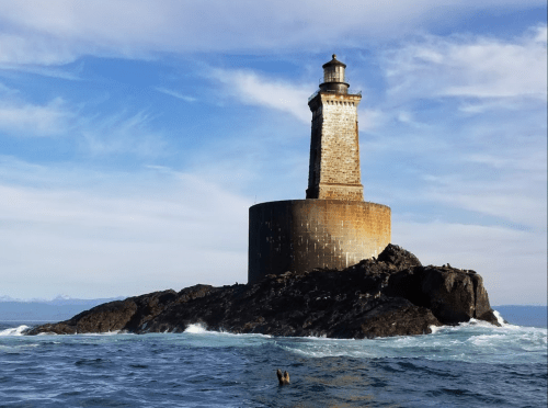 A tall lighthouse stands on a rocky island, surrounded by ocean waves under a clear blue sky.