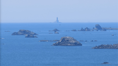 A distant lighthouse stands on a rocky island, surrounded by calm blue waters and scattered rocks in the foreground.