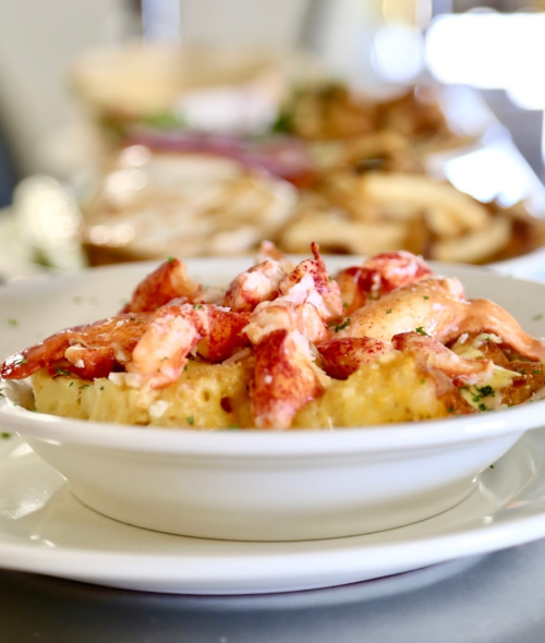 A bowl of creamy lobster mac and cheese topped with chunks of lobster and herbs, served in a restaurant setting.