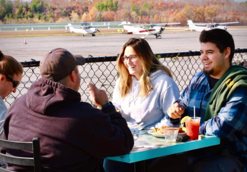A group of four friends enjoys a meal outdoors at an airport, with small planes in the background and autumn foliage.