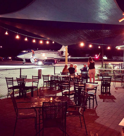 A nighttime scene at a restaurant patio with string lights, overlooking a private jet on the runway.