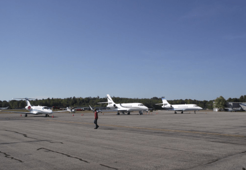 A busy airport tarmac with several private jets parked and a person walking nearby under a clear blue sky.