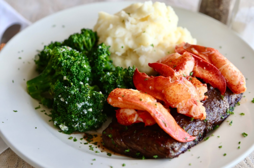 A plate featuring steak topped with lobster, served with broccoli and mashed potatoes, garnished with parsley.
