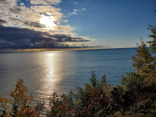 A serene view of a calm lake reflecting the sun, surrounded by trees and colorful autumn foliage.
