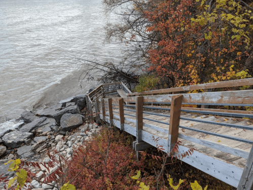 A wooden staircase leads down to a rocky shoreline, surrounded by autumn foliage and a calm body of water.