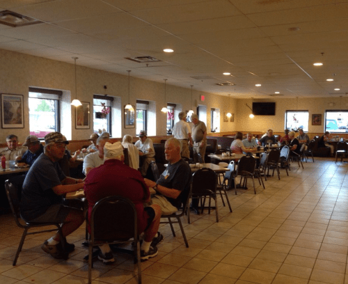A busy diner with groups of older men chatting and eating at tables, with warm lighting and a casual atmosphere.