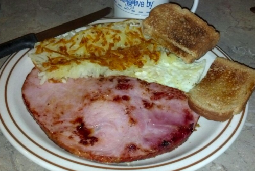 A plate with a slice of ham, scrambled eggs, hash browns, and two pieces of toast.