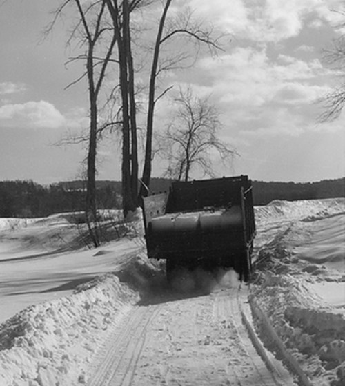 A truck navigates a snowy path surrounded by bare trees under a cloudy sky.