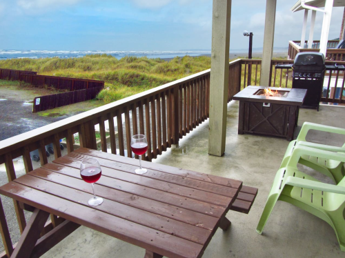 A cozy patio with two glasses of red wine, a fire pit, and a view of the ocean under a cloudy sky.