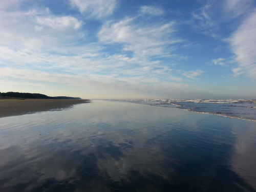 A serene beach scene with calm waters reflecting the sky and clouds, stretching into the horizon.