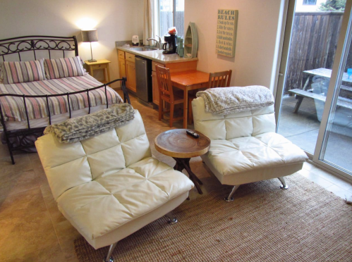 Cozy room with a bed, two white chairs, a wooden table, and a kitchenette, featuring warm lighting and a rug.
