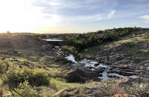 A serene landscape featuring a rocky terrain, a calm stream, and a distant dam under a bright sky.