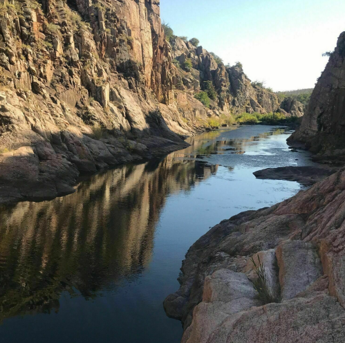 A serene river flows between rocky cliffs, reflecting the landscape under a clear blue sky.