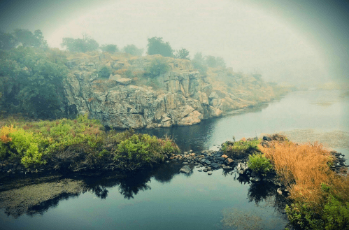 A misty landscape featuring a rocky cliff beside a calm river, surrounded by lush greenery and soft fog.