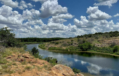 A serene river winds through rocky terrain under a bright blue sky filled with fluffy white clouds.