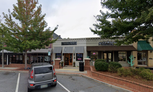 A street view of a shopping area featuring storefronts, trees, and parked cars.
