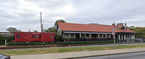 A red train car beside a restaurant with a sloped roof, set along a railway track under a cloudy sky.