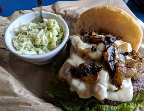 A juicy burger topped with grilled onions and mayonnaise, served with a side of coleslaw in a small bowl.