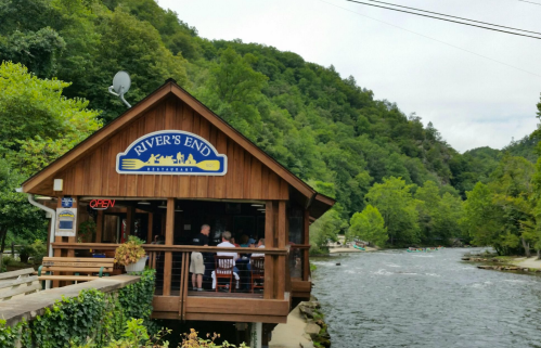 A wooden restaurant by a river, surrounded by green trees, with people dining on the patio. Sign reads "River's End."
