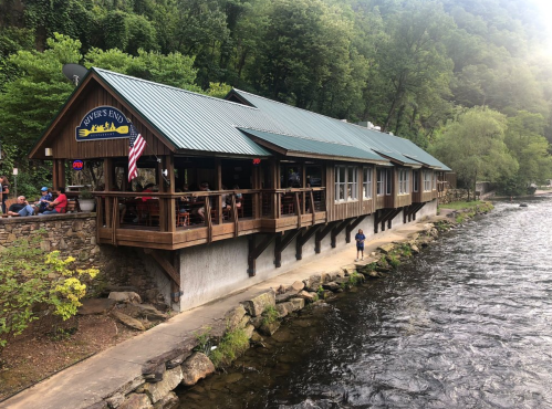 A riverside restaurant with a wooden deck, surrounded by greenery and people enjoying the outdoor seating.