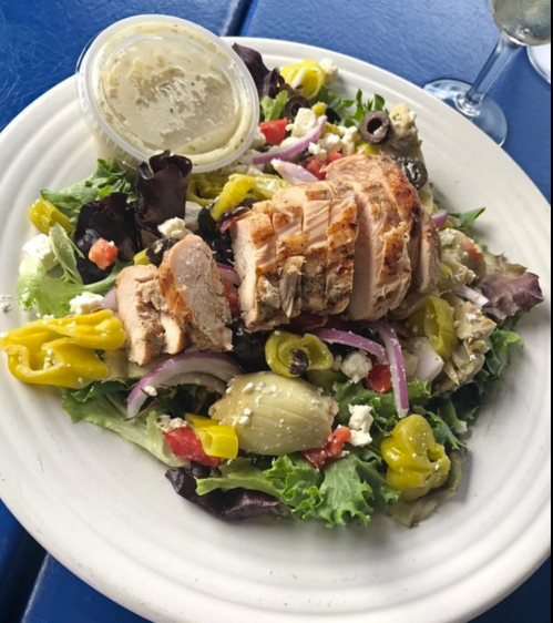 A colorful salad topped with grilled chicken, olives, peppers, and feta cheese, served with a dressing on the side.