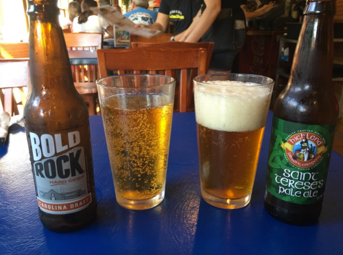 Three beverages on a blue table: a bottle of Bold Rock cider, a glass of golden beer, and a bottle of Saint Terese's pale ale.