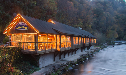 A cozy riverside restaurant with warm lights, surrounded by trees and autumn foliage, reflecting on the water.