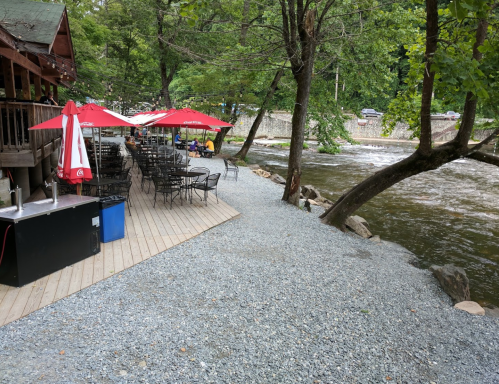 A riverside patio with tables and umbrellas, surrounded by trees and a flowing river.