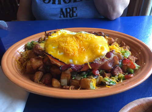 A colorful plate of breakfast hash topped with cheese, bacon, and a fried egg, served on a blue table.
