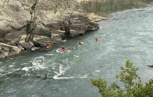 Three kayakers navigate a rocky river with swirling water, surrounded by steep cliffs and greenery.
