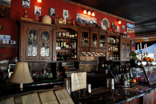 A rustic bar with wooden cabinets, various bottles, and framed photos on a red wall, creating a cozy atmosphere.
