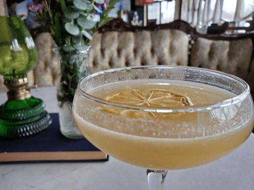 A close-up of a cocktail with a lemon slice, next to a vintage lamp and a vase of flowers on a table.