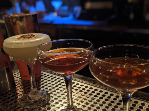 Three elegant cocktails on a bar counter, featuring a frothy drink with a dried citrus slice and two amber-colored drinks.