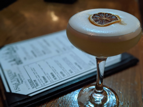 A cocktail with a frothy top and a dried lemon slice, placed next to a menu on a wooden table.
