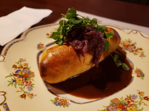A gourmet hot dog topped with greens, served on a decorative plate with a rich sauce.