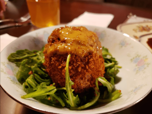 A crispy, golden-brown ball on a bed of greens, topped with a creamy sauce, served in a decorative bowl.
