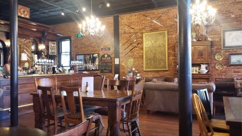 Cozy restaurant interior with wooden tables, a bar, brick walls, chandeliers, and decorative swords on display.