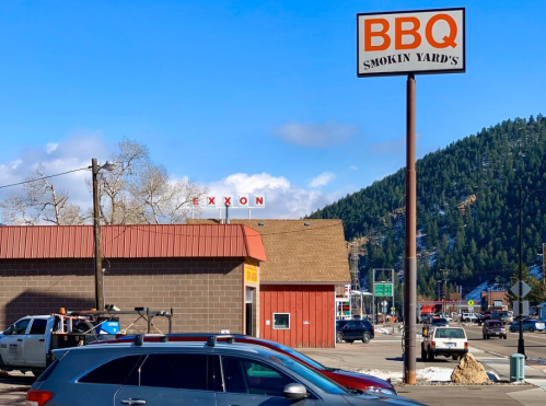 A BBQ restaurant sign next to an Exxon station, with mountains in the background and parked cars in the foreground.
