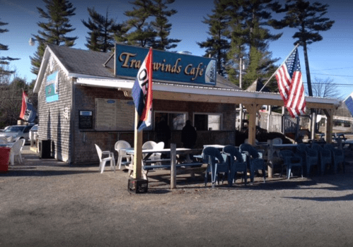A cozy café with outdoor seating, flags, and a sign reading "Tradewinds Café," surrounded by trees.