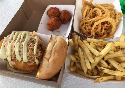 A meal featuring a pulled chicken sandwich with sauce, onion rings, fries, and three fried balls in a takeout box.