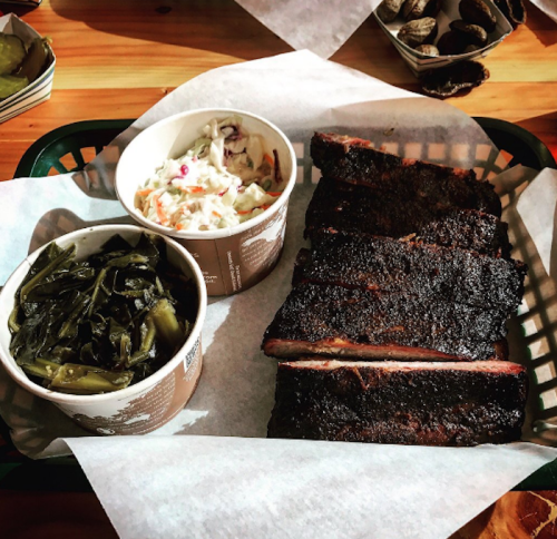 A basket of smoked ribs with sides of coleslaw and collard greens on a wooden table.