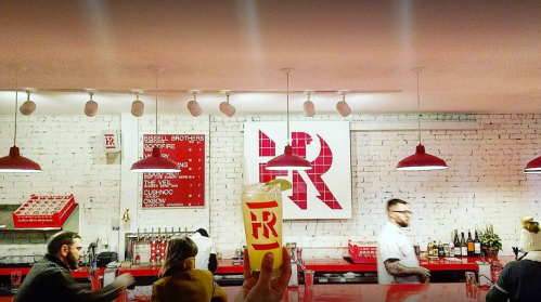 A bar scene with patrons, a person holding a drink, and a large red logo on the wall. Bright, modern decor.