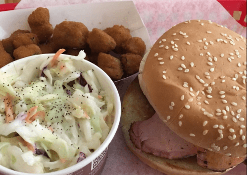 A plate featuring a sesame seed bun sandwich with sliced meat, coleslaw, and a side of fried okra.