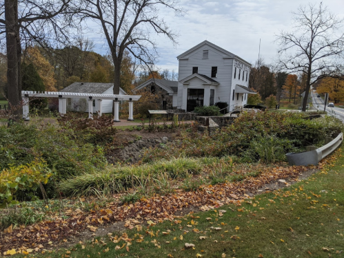A white house surrounded by greenery and autumn leaves, with a pathway and trees in the background.