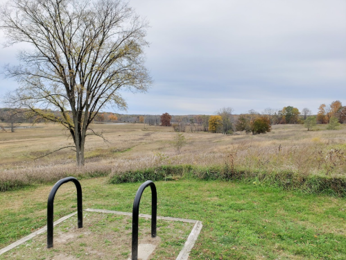 A grassy area with a bike rack, overlooking a vast landscape of fields and trees under a cloudy sky.