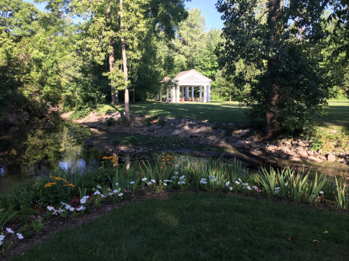 A serene landscape featuring a pond, colorful flowers, and a white gazebo surrounded by lush greenery.